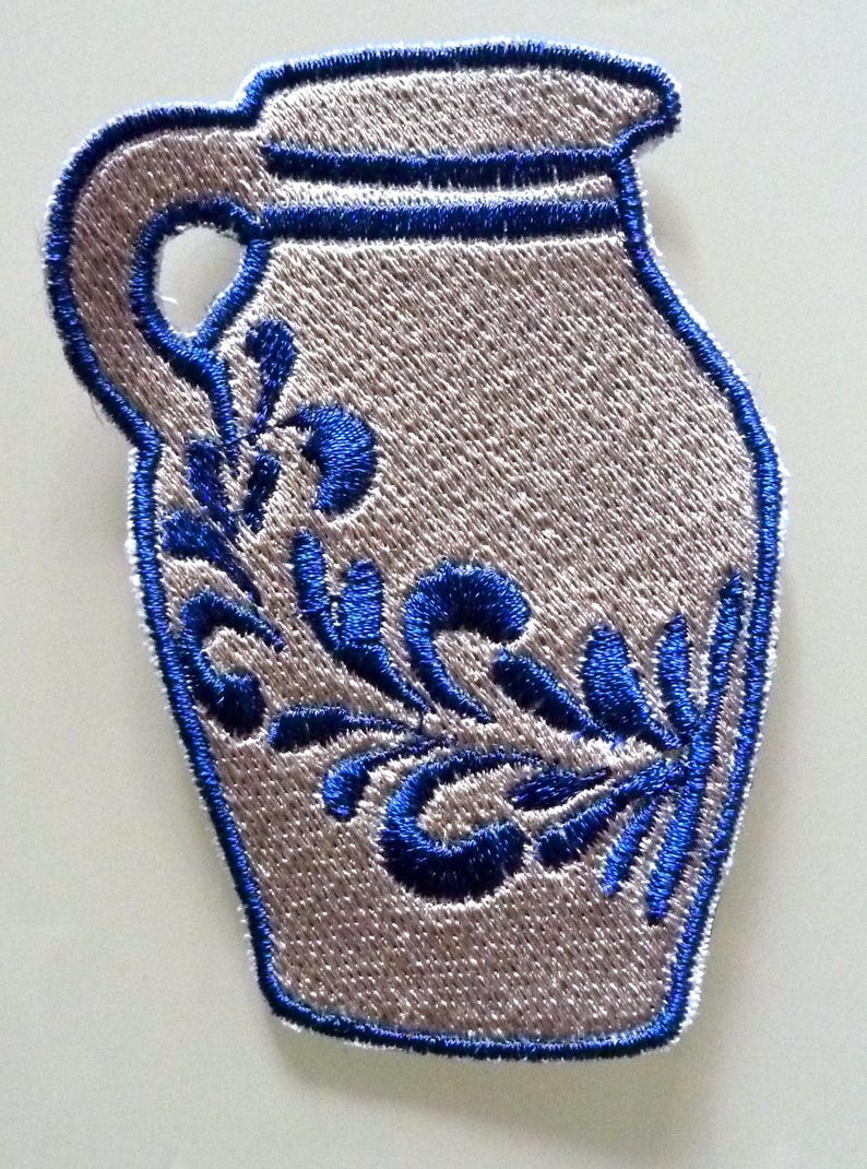 Puede incluir: Un parche bordado azul y blanco de una jarra de cer&aacute;mica con un dise&ntilde;o floral.