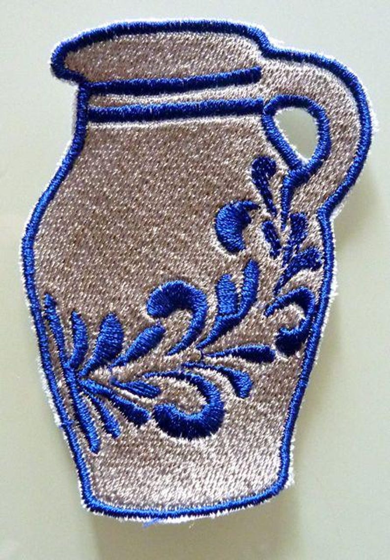 Puede incluir: Un parche de planchado beige bordado con un c&aacute;ntaro azul y blanco con un dise&ntilde;o floral.