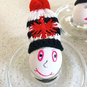 May include: A white egg decorated with a hand-drawn face featuring pink eyes and a smiling mouth, wearing a miniature knitted beanie. The beanie has a red pom-pom, white and black stripes, and a red eagle emblem on the front section.