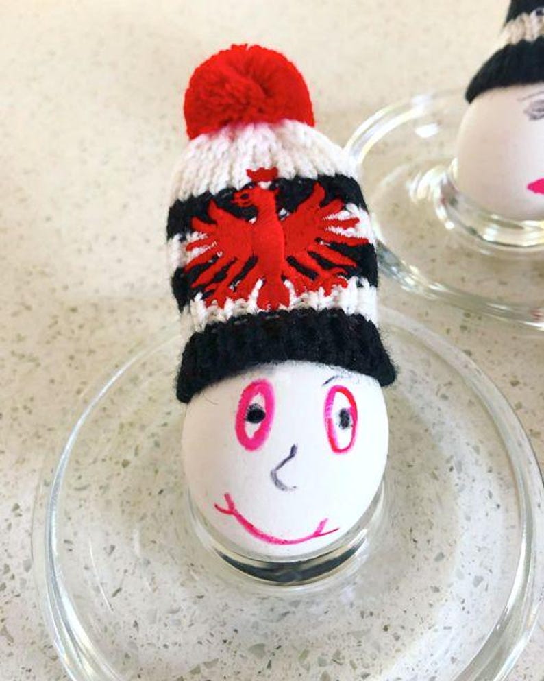 May include: A white egg decorated with a hand-drawn face featuring pink eyes and a smiling mouth, wearing a miniature knitted beanie. The beanie has a red pom-pom, white and black stripes, and a red eagle emblem on the front section.