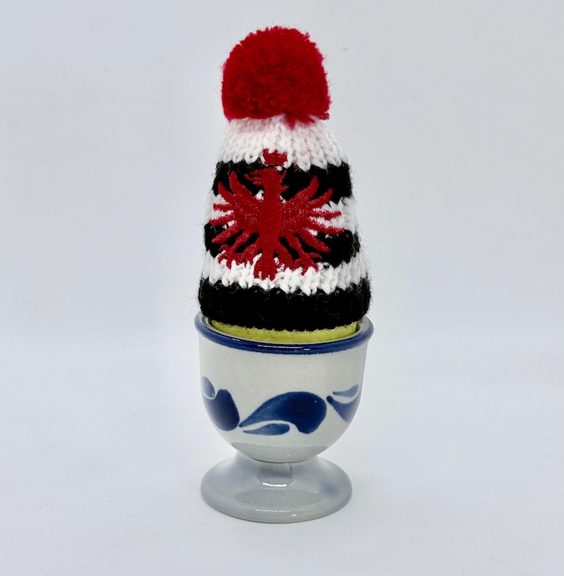 May include: A ceramic egg cup with a white base and blue painted leaf patterns, holding a yellow egg wearing a small knitted hat. The hat features horizontal white and black stripes, a red pom-pom on top, and a red eagle emblem on the front.