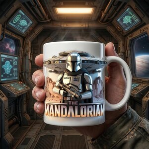 May include: White ceramic mug featuring a detailed image of The Mandalorian character with a blaster, a spaceship, and the text "THE MANDALORIAN". The mug has a curved handle and is held in a hand.