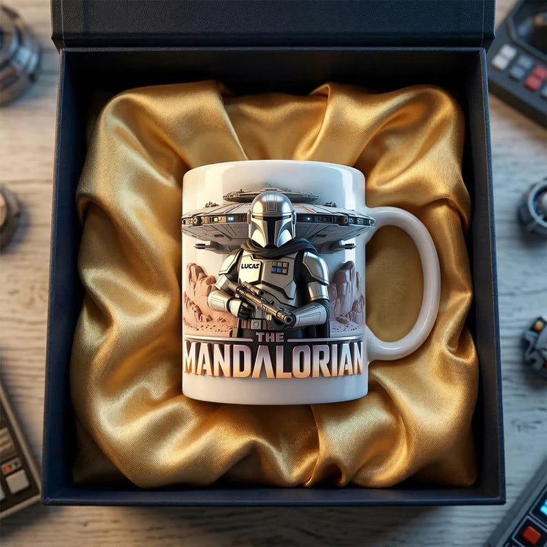 May include: White ceramic mug featuring The Mandalorian with a blaster, a spaceship, and the text "The Mandalorian." The mug is presented in a blue gift box with gold satin lining.