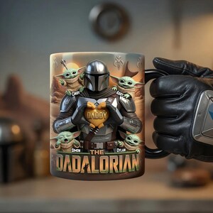 Puede incluir: Una taza de cer&aacute;mica con un dise&ntilde;o inspirado en The Mandalorian. La taza muestra al personaje de Mandalorian con figuras de Grogu y el texto "DADDY" y "THE DADALORIAN". La taza es sostenida por una mano enguantada.