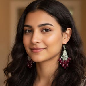 Peut inclure: Une personne aux cheveux noirs et aux yeux bruns portant des boucles d'oreilles en argent. Chaque boucle d'oreille présente un groupe de petites perles rondes rouge foncé suspendues à une chaîne en argent.