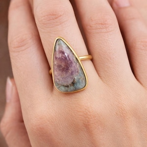 Könnte beinhalten: Ein goldener Ring mit einem großen, unregelmäßig geformten Edelstein in Lila-, Rosa- und Grüntönen. Der Stein ist in eine goldene Lünette gefasst. Das Band des Rings ist schlicht, poliertes Gold.