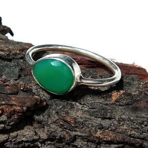 Könnte beinhalten: Ein silberner Ring mit einem leuchtend grünen Edelstein. Der oval geformte Stein ist in einem schlichten, eleganten Band gefasst. Das Design des Rings betont die Farbe und Form des Edelsteins.
