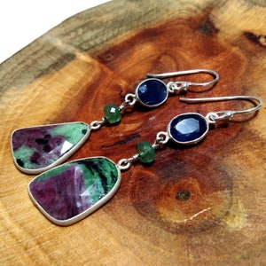 Puede incluir: Pendientes colgantes con piedras preciosas de rubí y zafiro. Los pendientes tienen un engaste plateado y un cierre de gancho. Los rubíes son rojos y verdes, mientras que los zafiros son de color azul oscuro. Se muestran sobre una superficie de madera.