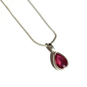 Op de afbeelding: Een zilveren ketting met een druppelvormige hanger. De hanger heeft een dieprode edelsteen gezet in een zilveren bezel. De ketting is een dunne slangenketting, op een witte achtergrond.