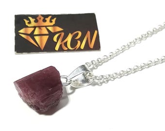 May include: A silver chain necklace with a raw, dark pink gemstone pendant. The pendant is a rough, rectangular shape. A black business card with gold text and a diamond logo is in the background.
