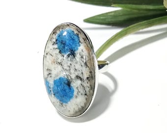 May include: Silver ring with an oval k2 stone. The stone has a white base with grey and black speckles and two bright blue spots. The ring is set against a white background with green foliage.