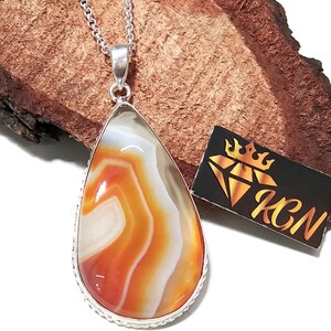 Puede incluir: Un colgante en forma de lágrima con una piedra de ágata bandeada con capas de color naranja, blanco y gris translúcido. La piedra está engastada en un marco de metal plateado y cuelga de una cadena plateada. Se ve una etiqueta negra con una corona dorada y las letras KGN.