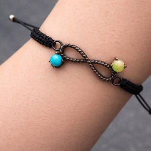 May include: Black cord bracelet featuring an infinity symbol design. The bracelet has a turquoise and a yellow-green gemstone. The infinity symbol is made of a dark metal. The bracelet is adjustable.