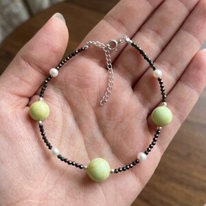 Può includere: Un delicato braccialetto con tre perline sferiche verde pallido intervallate da piccole perline nere e perle bianche. Il braccialetto ha una catena e una chiusura argentate ed è esposto su una mano.