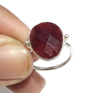 May include: A silver ring featuring a large, oval-shaped, faceted deep red gemstone set in a smooth metal bezel. The band is thin and polished, with small decorative metal spheres attached to the sides where the setting meets the ring band.
