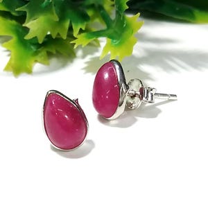 May include: A pair of teardrop-shaped stud earrings. Each earring has a polished, magenta-coloured gemstone set in a silver-coloured metal bezel. The earrings are displayed on a white surface with green foliage in the background.