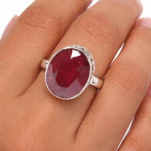 Può includere: Un anello d'argento con un grande rubino ovale. Il rubino è di un colore rosso intenso ed è incastonato in una fascia d'argento con un motivo decorativo a filigrana attorno alla pietra. L'anello è mostrato su un dito.