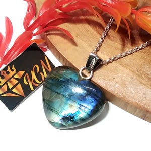 Puede incluir: Colgante de labradorita en forma de corazón con cadena de plata. La piedra preciosa muestra tonos iridiscentes azules y verdes. El colgante se exhibe sobre una superficie de madera con acentos florales rojos y naranjas. Una etiqueta negra con texto dorado es visible.