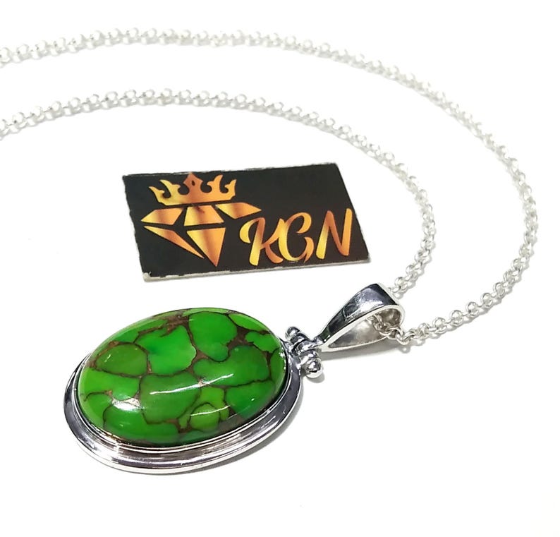 Puede incluir: Un collar de cadena de plata con un colgante ovalado verde con una piedra turquesa. El colgante tiene un borde de plata y una argolla de plata.