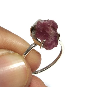 Puede incluir: Un anillo de plata con una gema de rubí en bruto. El rubí es de un color rojo rosado intenso con una superficie rugosa y texturizada. La banda del anillo es de plata pulida simple.