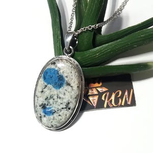 Puede incluir: Collar con colgante ovalado con una piedra K2 con patrones azules y blancos, engastada en un marco de metal plateado. El colgante cuelga de una delicada cadena plateada. El collar se muestra sobre un fondo verde.