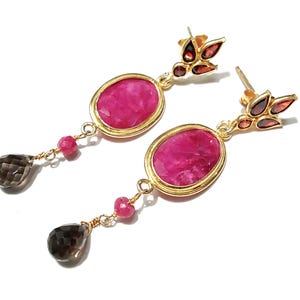 May include: A pair of gold earrings featuring oval, magenta gemstones. The earrings have smaller pink and brown gemstones, and a leaf design with red gemstones. The earrings are set against a white background.