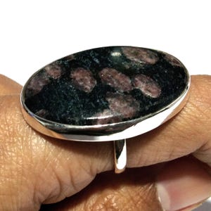 May include: A ring featuring a large, polished, oval-shaped stone with a dark black base and scattered, rounded maroon-colored inclusions. The stone is set within a smooth, polished silver-toned metal bezel and band, creating a bold statement piece.