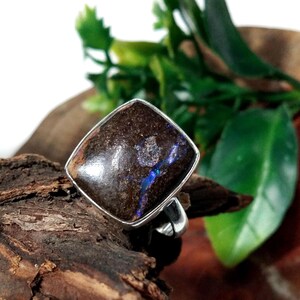 Pode incluir: Um anel de prata com uma pedra preciosa opala boulder de formato quadrado. A pedra tem uma superfície texturizada castanho-escura com um veio iridescente azul vibrante que atravessa o centro. O aro é de prata lisa e polida.