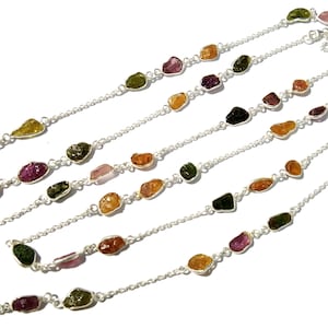 Può includere: Diverse collane con pietre preziose grezze e colorate infilate su delicate catene d'argento. Le pietre sono di vari colori, tra cui verde, rosa, arancione e giallo. Sfondo bianco.