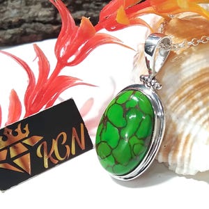 Puede incluir: Un colgante de plata con una piedra turquesa verde engastada en un marco ovalado de plata. El colgante est&aacute; sobre una concha blanca.