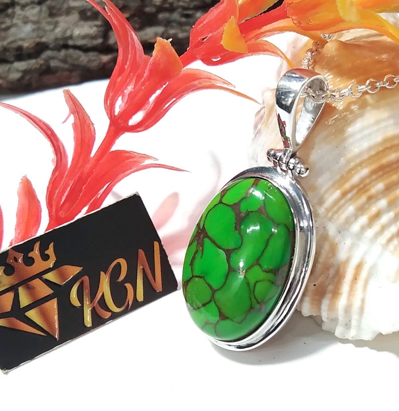 Puede incluir: Un colgante de plata con una piedra turquesa verde engastada en un marco ovalado de plata. El colgante est&aacute; sobre una concha blanca.