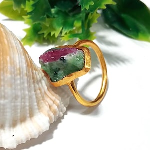 Könnte beinhalten: Ein goldfarbener Ring mit einem rohen Edelstein in Grün- und Rottönen. Der Edelstein ist in einer rechteckigen Fassung gefasst. Der Ring wird auf einer Muschel vor weißem Hintergrund präsentiert.