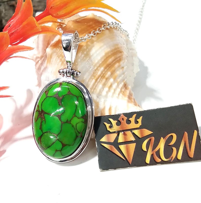 Puede incluir: Un collar con un colgante de plata adornado con una piedra turquesa ovalada verde. La piedra presenta un patr&oacute;n de m&aacute;rmol con vetas marrones. El colgante est&aacute; en una cadena de plata y se muestra en una concha de mar.