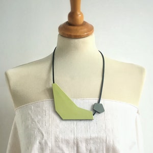 Puede incluir: Un collar geométrico con un colgante grande de color verde lima y un colgante más pequeño de color gris. El collar está hecho de cordón negro y se lleva sobre un maniquí blanco.