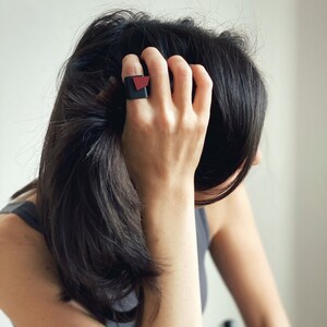 Puede incluir: Un anillo geométrico negro y rojo. El anillo presenta una base cuadrada negra con un acento angular rojo. El anillo se lleva en un dedo, con la mano y el brazo visibles. El fondo es de un color suave y neutro.