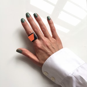 Könnte beinhalten: Ein geometrischer Ring in Schwarz und Orange mit quadratischer Form. Der Ring wird am Zeigefinger einer Hand mit grünem Nagellack getragen.
