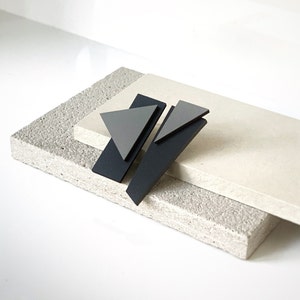 Puede incluir: Un par de pendientes geométricos con triángulos negros y grises. Los pendientes están hechos de un material ligero y tienen un diseño moderno y minimalista.