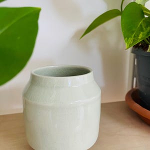 Pode incluir: Um vaso de cerâmica verde claro com acabamento craquelado. O vaso cilíndrico tem uma base arredondada e uma borda ligeiramente alargada. Está sobre uma prateleira de madeira clara, com plantas verdes ao fundo.