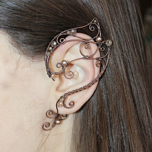 May include: A copper wire ear cuff with a delicate, intricate design. The cuff is adorned with small, clear and pearl-colored beads. The cuff is worn on the upper part of the ear, extending from the helix to the top of the ear.