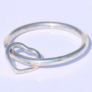 Könnte beinhalten: Ein silberner Ring mit einem herzförmigen Anhänger. Das Ringband ist ein einfacher, glatter Kreis, und der Herzanhänger ist am Band befestigt. Der Ring ist wahrscheinlich aus Sterlingsilber oder einem ähnlichen Metall gefertigt.