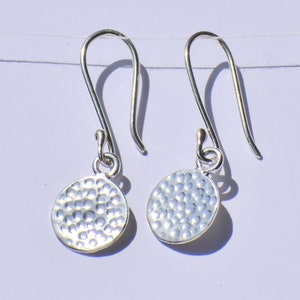 May include: A pair of silver drop earrings. Each earring features a hammered, circular charm suspended from a curved ear wire. The charms have a textured surface with a dimpled pattern.
