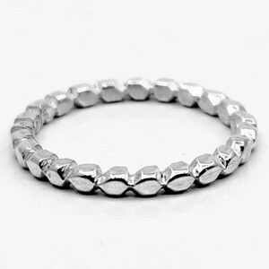 Op de afbeelding: Een zilveren ring met een herhalend geometrisch patroon. De ring heeft een reeks kleine, facetvormige vormen die rond de band gaan. De ring staat tegen een witte achtergrond, wat de metaalglans en het ingewikkelde patroon benadrukt.
