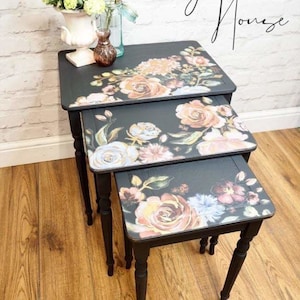 Può includere: Un set di tre tavolini a incastro neri con motivi floreali. I tavolini sono dipinti di nero e presentano un motivo floreale in tonalità di rosa, blu e verde.