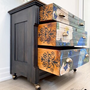 Op de afbeelding: Een zwart geverfde houten dressoir met vier lades. De lades zijn versierd met een bloemenpatroon en een damastontwerp. Het dressoir heeft messing handgrepen en staat op vier zwarte wielen.