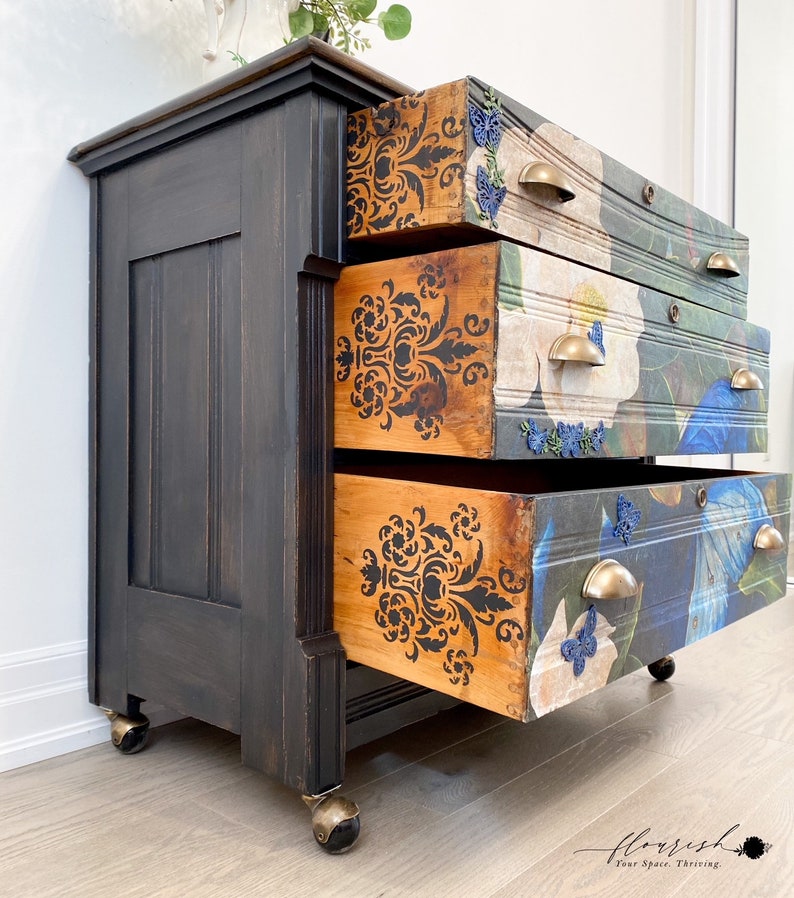 Op de afbeelding: Een zwart geverfde houten dressoir met vier lades. De lades zijn versierd met een bloemenpatroon en een damastontwerp. Het dressoir heeft messing handgrepen en staat op vier zwarte wielen.
