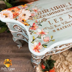 Può includere: Un tavolo dipinto di azzurro con dettagli dorati e un motivo floreale sul piano. Il motivo floreale presenta fiori rosa, arancioni e bianchi. Il tavolo ha un piano curvo e tre gambe. Il testo sul tavolo recita "LIBRAIRIE L GÉRARDMER (Vos) Compte Postal Lyc LES A 1, Rue Villars- M-MATHIEU".