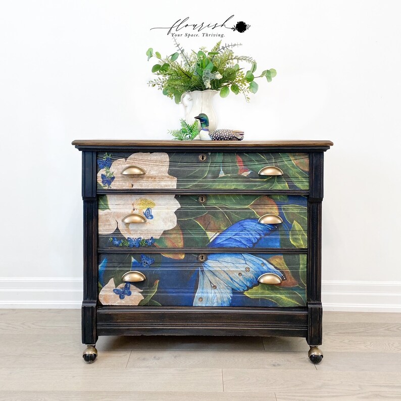 Op de afbeelding: Een zwart geverfde commode met drie laden heeft een bloemen- en vlinderontwerp. De laden hebben messing knoppen. De commode staat op een houten vloer.