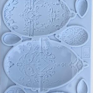 Op de afbeelding: Een lichtblauwe siliconen mal voor het maken van sierlijke eieren. De mal heeft gedetailleerde, sierlijke ontwerpen en bevat de tekst "Silikon Formen" en "Silicone Mould". De verpakking is gelabeld "LABLANCHE" en meet 12,7 x 20,3 cm.