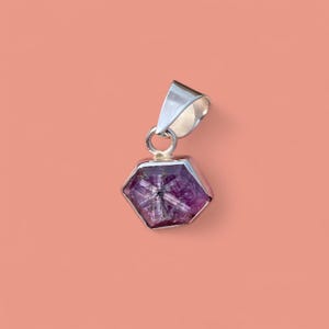 May include: A hexagonal pendant with a silver bail and setting. The pendant features a faceted, deep purple gemstone with a star-like pattern. The background is a solid peach color.