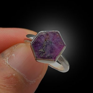 以下が含まれることがあります： 六角形の濃いマゼンタ色の宝石をあしらったシルバーリング。宝石は、内部の線とインクルージョンを持つテクスチャード加工された表面を持っています。リングは暗い背景に対して保持され、石の鮮やかな色を強調しています。